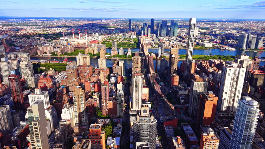 Queensboro bridge spans East river toward Manhattan skyline, New York City. Roosevelt island greenery lines the water beneath sunny sky.