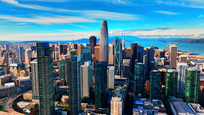 Downtown San Francisco skyline with skyscrapers. Overhead view of glass high rise buildings, freeway ramps and distant hills under bright blue sky.