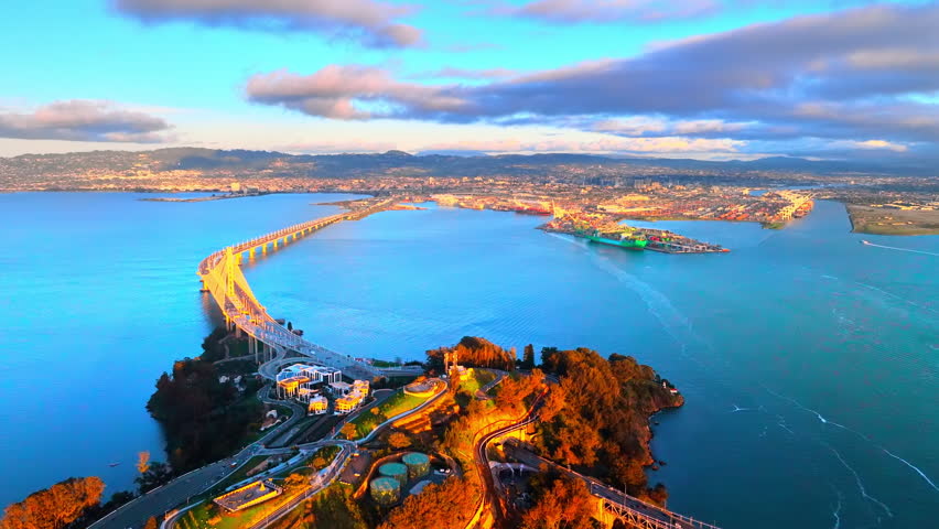 Sunset over the Bay Bridge and Oakland port, San Francisco, California. Aerial view of Treasure Island, container cranes and cargo ships along the shoreline at dusk.