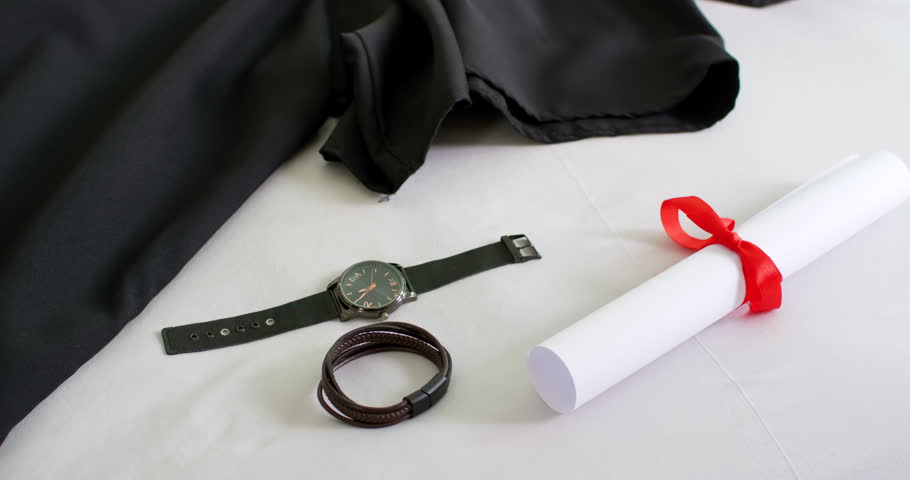 Camera panning zooming revealing black gown, watch, bracelet, diploma on white fabric for grad prep. Robe, graduation, scroll, ribbon, timepiece, leather, stilllife