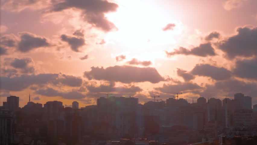 Timelapse: setting sun shining through fast moving clouds over Istanbul before hiding behind silhouettes of buildings and cranes. The city gets darker under orange sunset sky - time lapse