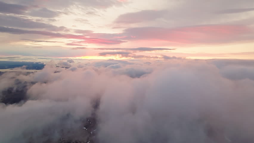 Aerial view above the clouds at sunrise over Iceland’s Highlands as colorful morning light illuminates dramatic cloud layers revealing glimpses of volcanic terrain below.