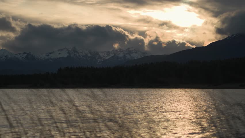The view of a mountain range with snow-capped peaks seen over a calm lake through foreground grass at sunset.
