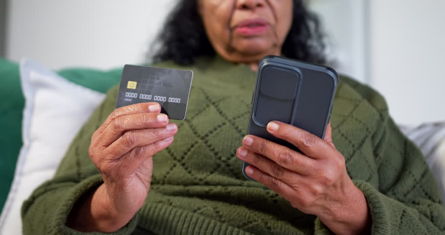 Senior African woman sitting in green knit on couch holding credit card and phone verifying payment. Elderly, home, mobile, touchscreen, fintech, modern, focused