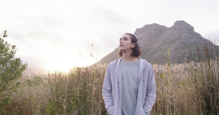 Man in his 20s standing in grassy field at foothill, camera closing in, turning head, smiling. Silhouette, backlit, meadow, goldenhour, hoodie, casual, nature