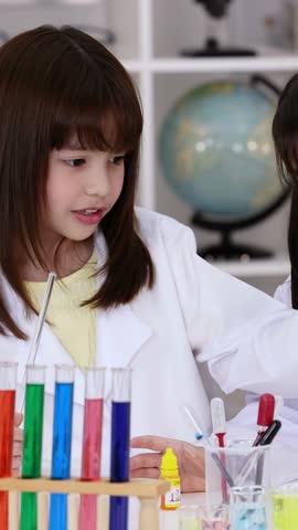 Two young Asian girls wear lab coats while mixing colorful liquids in a classroom setting.