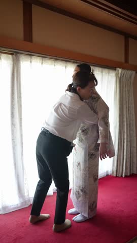 Woman assists another in adjusting her kimono. Soft light fills the traditional room. The helper focuses on fitting the garment. The recipient smiles gently, standing still. Perfect for cultural