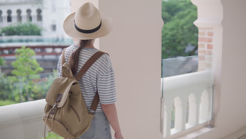 Female tourist in hat with backpack is walking along scenic galleria inside the Sultan Abdul Samad Building and enjoying city view in Kuala Lumpur, Malaysia. Awesome interior of the colonial building.