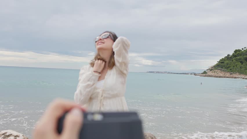 point of view perspective of a photographer taking pictures of a beautiful young woman posing on a sandy beach. The model enjoys the photo session, smiling and moving naturally by the sea