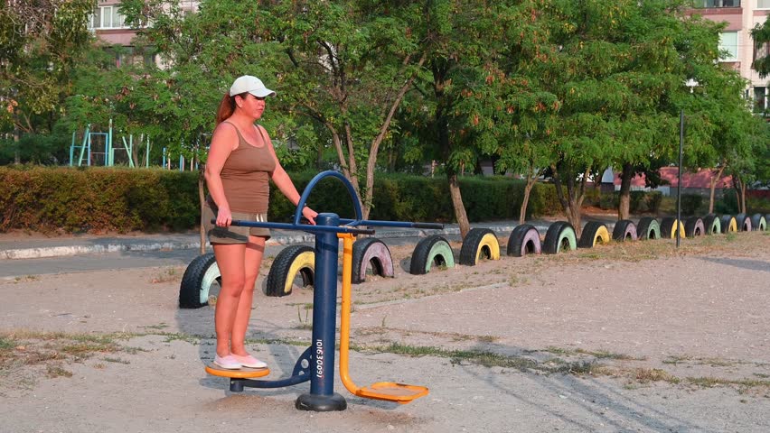 woman wearing a cap training legs and coordination on a blue and yellow outdoor stepper machine at a sports playground