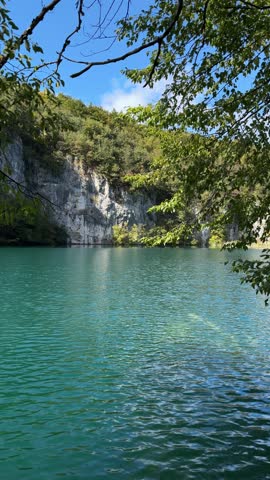 Plitvice Lakes National Park, Croatia. Serene lake view with lush green reeds reflecting in calm water, surrounded by forested hills under clear blue sky
