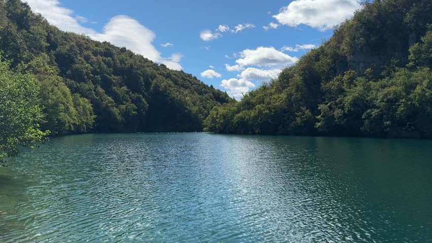 Plitvice Lakes National Park, Croatia. Serene lake view with lush green reeds reflecting in calm water, surrounded by forested hills under clear blue sky