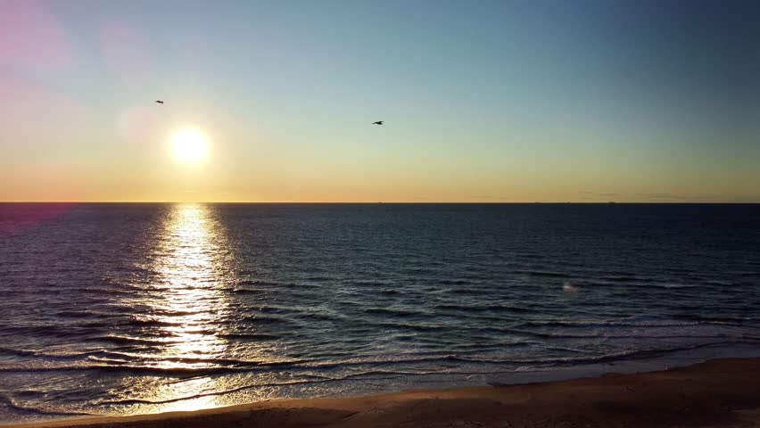 Aerial drone footage captures a stunning and tranquil summer sunset over the North Sea at Hirtshals, Denmark. The low sun hangs just above the horizon, its golden light creating a glittering path across the surface of the calm water. Gentle waves roll onto the sandy beach in the foreground. The sky is a clear, beautiful gradient of orange, yellow, and blue. In the far distance, the silhouettes of several ships are visible on the horizon, adding to the vast maritime scenery of the North Jutland coast.