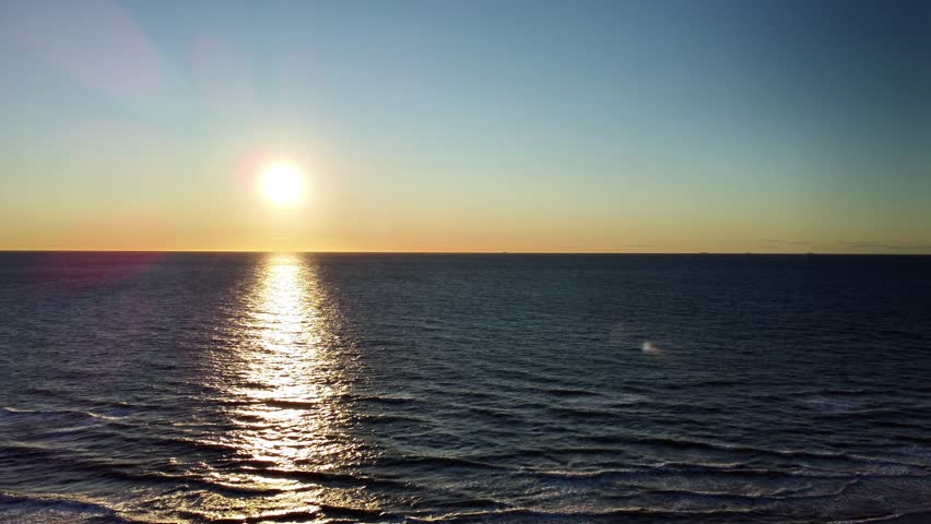 Aerial drone footage captures a breathtaking sunset over the open sea off the coast of Hirtshals, Denmark. The sun hangs low on the horizon, casting a brilliant golden reflection that shimmers across the surface of the dark blue water, likely the North Sea or Skagerrak strait. The sky transitions from a deep blue to a warm, clear orange and yellow at the skyline. The calm waves gently ripple, creating a serene and peaceful atmosphere. This scenic video showcases the vast, natural beauty of the Danish coastline during a tranquil summer evening.