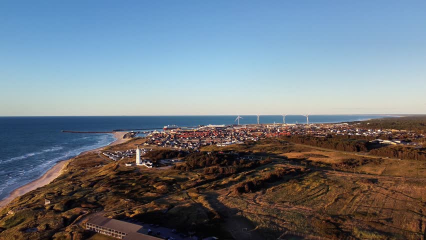 Stunning aerial drone footage capturing the picturesque coastal town of Hirtshals in North Jutland, Denmark. The camera offers a wide panoramic view of the town, its bustling harbor with a large ferry, and the iconic white Hirtshals Lighthouse (Hirtshals Fyr) standing prominently on the dunes. The scene is bathed in the warm, golden light of a late summer afternoon under a clear blue sky. In the background, modern wind turbines line the horizon, showcasing a blend of tradition and renewable energy. The waves of the North Sea gently roll onto the sandy beach.