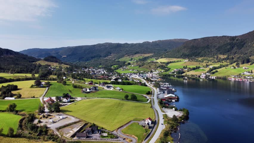 Stunning aerial drone footage capturing the serene beauty of a small village on the coast of the Vindafjord fjord in Rogaland, Norway. The camera provides a wide, panoramic view of the picturesque settlement, with its charming houses and farms scattered across lush green fields and rolling hills. A coastal road follows the shoreline of the deep blue fjord, where a small marina is visible. The scene is bathed in the bright sunlight of a beautiful late summer day, under a clear blue sky, showcasing the idyllic and tranquil Norwegian countryside.