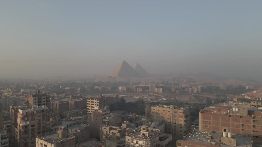 Aerial drone footage moving forward and upward from a dense urban Cairo neighborhood toward the Pyramids of Giza in the distance, showing the contrast between the ancient monuments and the modern city, Egypt.