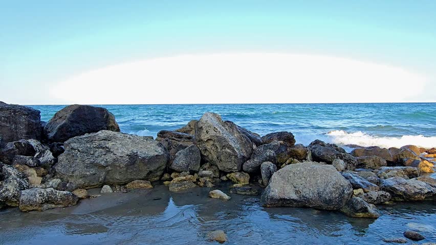 Weathered rocks stretch along the shoreline as calm turquoise waves ripple gently around them, the rocks catching warm golden light from the setting sun, the rocks creating a rugged natural coastline that contrasts beautifully with the soft evening sky