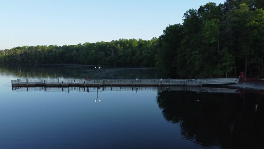 Serene aerial drone footage of Burke Lake at dusk. A long fishing pier extends from a densely forested, lush green shoreline into dark, tranquil waters. A few people stand on the illuminated dock enjoying the peaceful early morning atmosphere, with faint mist rising off the water. This picturesque, low-light landscape is perfect for nature documentaries, outdoor recreation promos, state park tourism, and cinematic establishing shots needing a quiet, reflective, and scenic environment.
