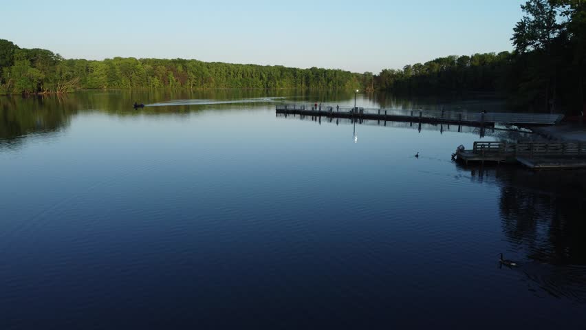 Aerial drone footage of tranquil Burke Lake, VA on a clear day. A long fishing pier extends into the calm blue water where people are fishing and relaxing. A small boat motors across the lake, leaving a gentle wake, while a lush green forest lines the distant shore. A lone goose swims gracefully in the foreground. This scenic video is ideal for projects highlighting outdoor recreation, nature, state parks, fishing, and peaceful summer landscapes.