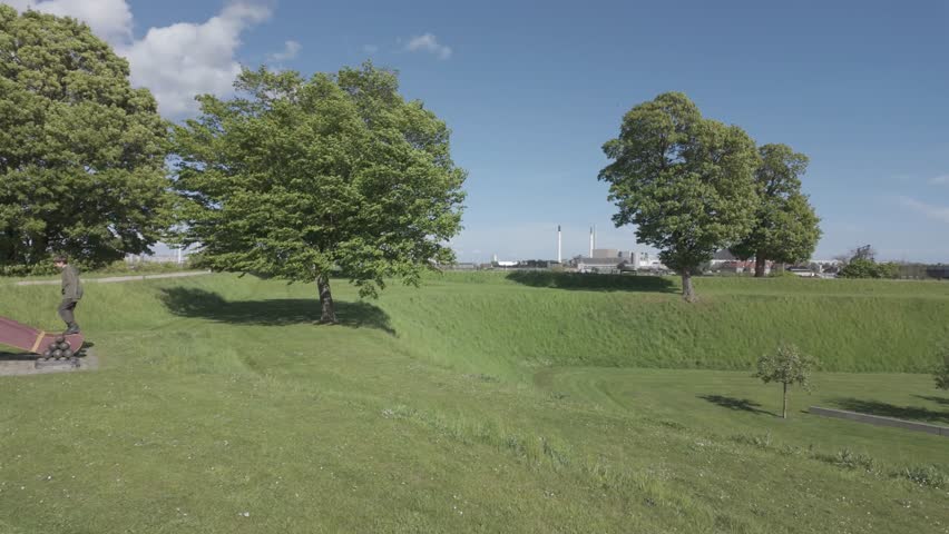 A historic cannon stands on the green earthwork rampart of Kastellet citadel beneath a spreading oak tree under a blue spring sky.