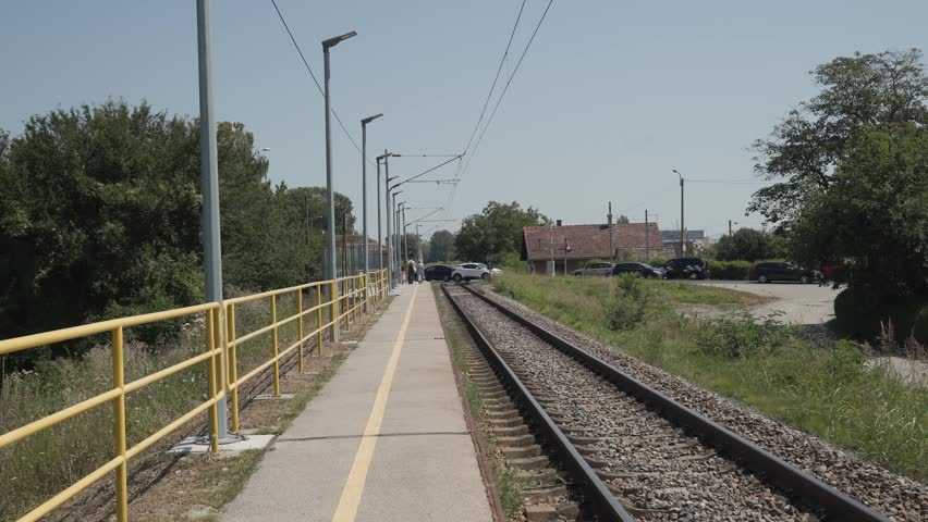 A small suburban rail platform at Remetinec railway station stretches alongside train tracks lined by overhead wires and greenery, with a distant train approaching under a clear blue sky.