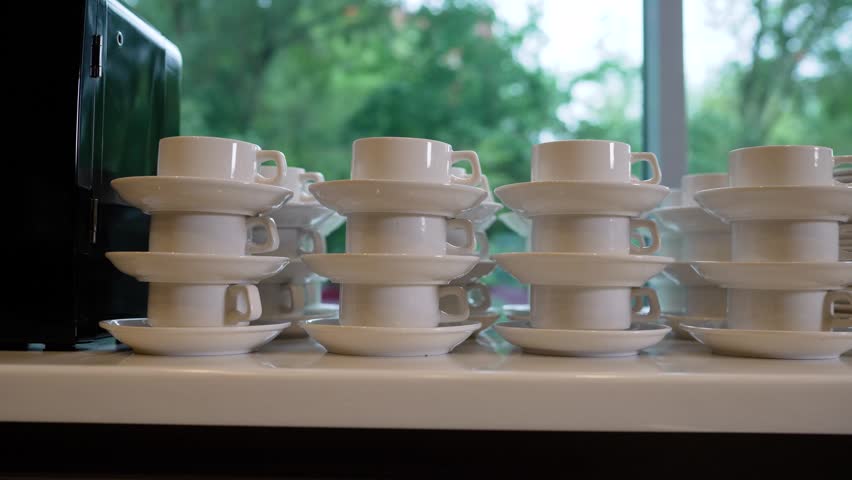 Multiple white porcelain cups saucers stacked in neat rows on glossy table. Soft natural light from window creates gentle reflections. Minimalist composition with clean lines neutral tones