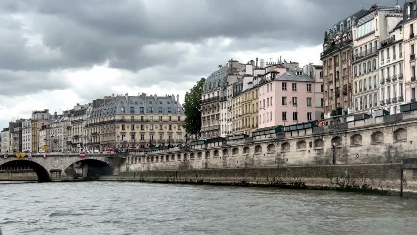 Scenic view of the historic Parisian architecture along the banks of the Seine River in Paris, France, featuring traditional European apartment buildings, stone bridges, and calm water under a dramatic overcast sky during a daytime boat cruise.