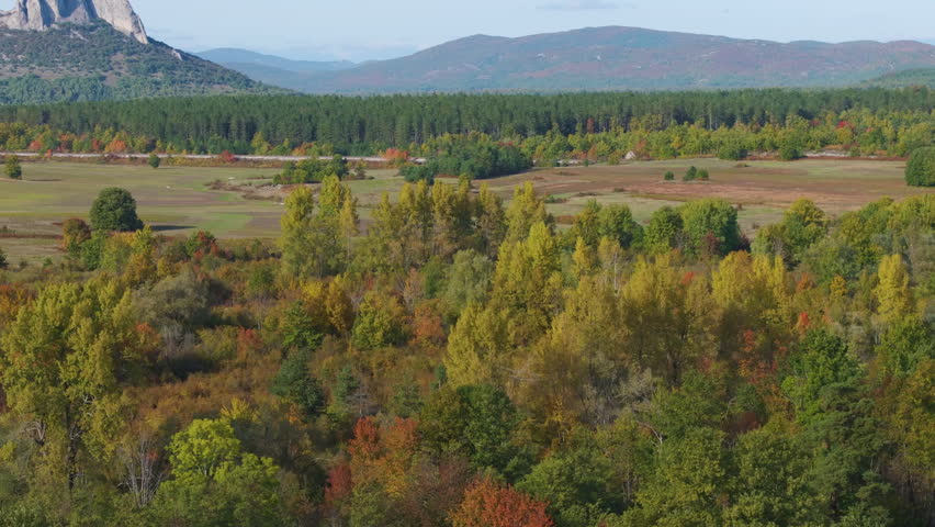 AERIAL: Rocky peak overlooking an autumn valley near Otočac, Croatia. Colorful woodland, open pasture, farmland and distant hills form a layered rural panorama beneath a pale blue sky.