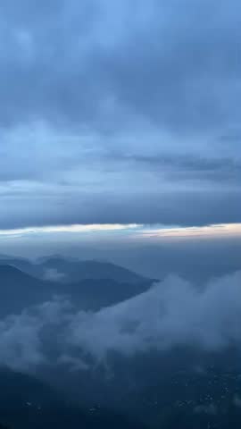 Dramatic blue hour sky over misty mountains with clouds flowing through green valleys at dawn. Soft orange glow on the horizon above fog. Peaceful and serene nature landscape from Pakistan mountains