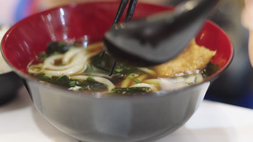 Close-up of chopsticks lifting thick udon noodles and seaweed from a bowl of hot Japanese soup. Professional food photography highlighting traditional Asian cuisine and authentic dining experience.