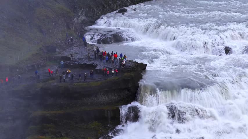 Aerial view showing diverse tourists gathered on dark basalt rock ledge observing massive rushing Gullfoss waterfall. Cinematic footage capturing powerful water currents and misty landscape.