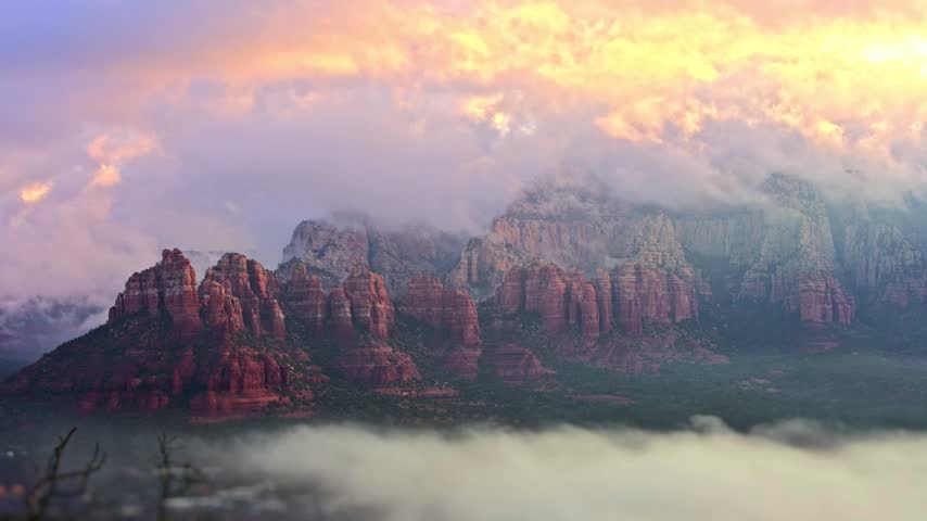 Breathtaking landscape cinematography capturing majestic red rock mountain peaks hidden behind dense moving mist under soft golden sunrise clouds in beautiful Sedona Arizona wilderness area.