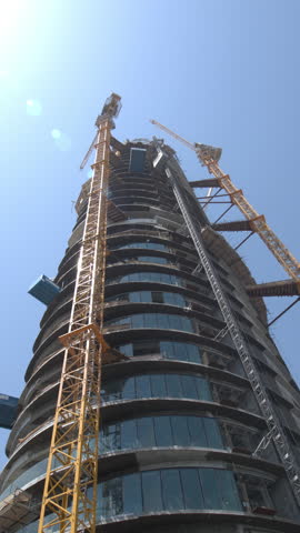 A tall circular skyscraper under construction in Dubai with large yellow tower cranes surrounding the building framework against a clear sky