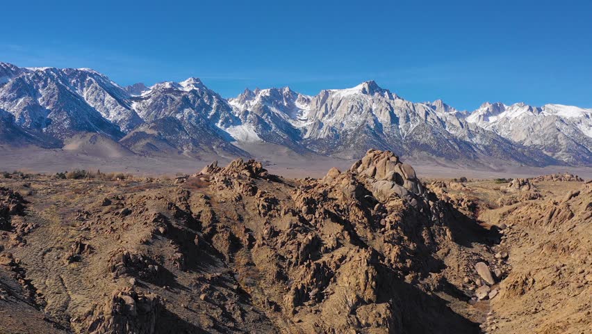 Aerial view showcasing rugged desert landscape featuring sharp granite boulders in foreground and magnificent snow covered peaks of Sierra Nevada mountain range under clear blue sky.