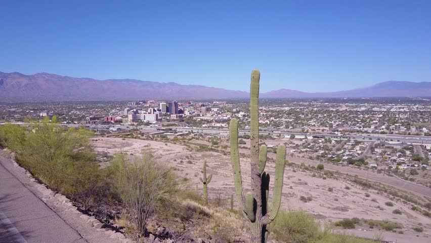 Aerial cinematic footage captures sprawling cityscape of Tucson Arizona set against mountain range with desert landscape and saguaro cacti in foreground during clear sunny day.