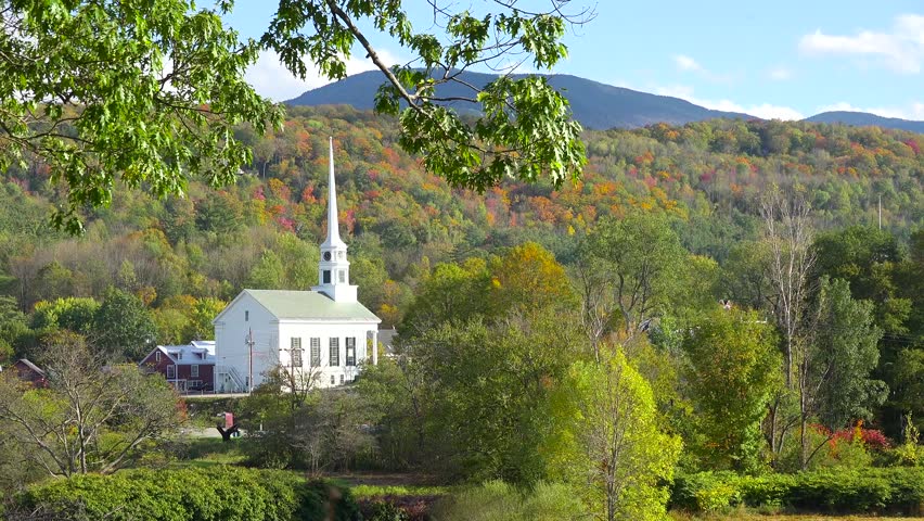 Footage showcases the white steeple of the historic first church nestled in a valley, framed by lush green branches. A colorful forest and mountains provide a picturesque autumnal background.