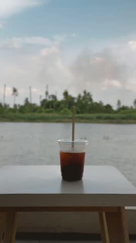 Iced coffee in a clear plastic cup with a straw on a white table. A calm river and lush green trees are visible.