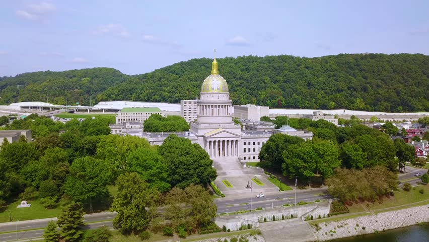 Aerial view of West Virginia state capitol building with gold dome in Charleston, featuring surrounding greenery, Kanawha river, and city infrastructure under bright blue sky.