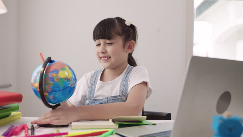 Cute Asian schoolgirl sitting at a desk using laptop computer for studying online learning via video calling and a remote teacher while having a geography lesson with a globe model in homeschool room. 