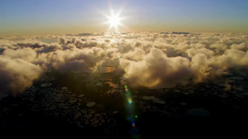 Stunning aerial view flying above fluffy clouds toward a bright sun flare during golden hour sunset over landscape below.