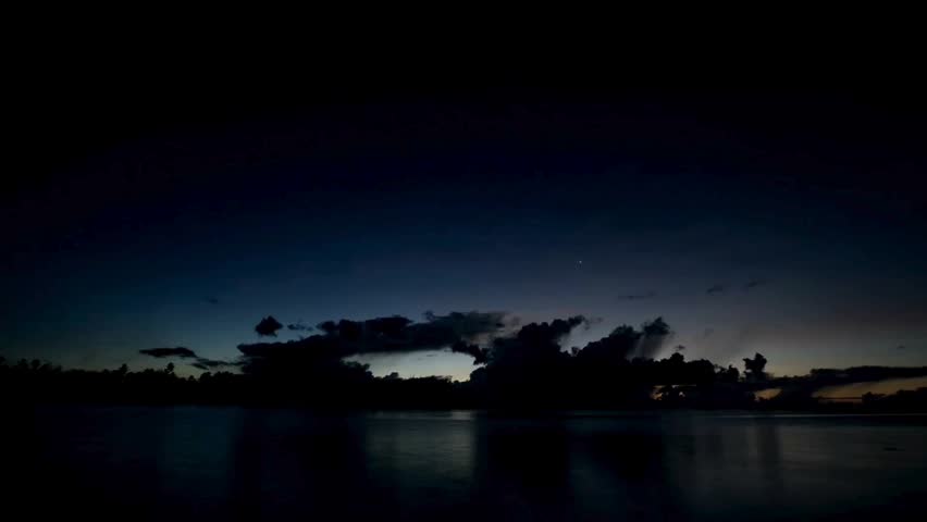 Beautiful night to day time lapse showing dramatic clouds, sunrise over calm ocean water, and palm tree coastline silhouette