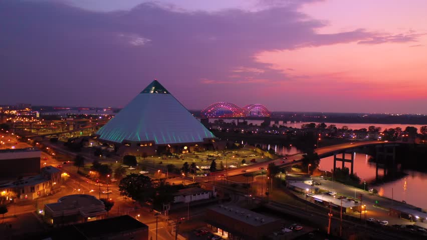 Aerial view of Memphis Pyramid illuminated at sunset over Mississippi river showcasing urban architecture city lights transportation infrastructure and scenic twilight horizon in Memphis.