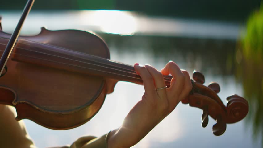 Close-up of a violinist playing soothing classical music outdoors by a tranquil lakeside at sunset, highlighting delicate finger placement and natural reflections