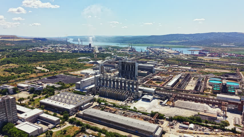 Factories and buildings are spread out near a river. There are green areas in the foreground. Smoke rises from chimneys. The scene shows a busy industrial landscape in bright sunlight.
