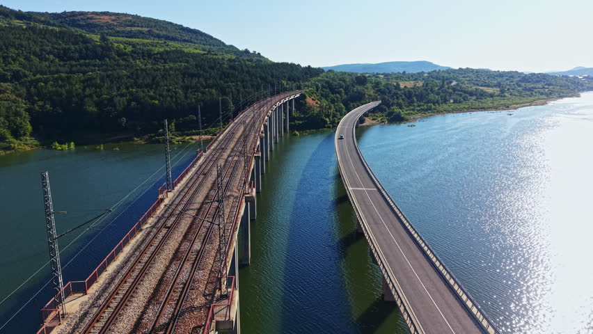 A railway bridge and a highway bridge cross over a body of water surrounded by hills and trees. The sun shines brightly over the scene, and a car travels on the road.
