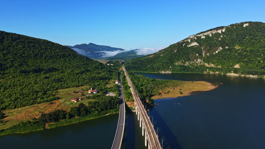 A bridge stretches across a river with hills and trees on both sides. The sky is clear and blue. There are small houses near the water. Mountains rise in the distance.