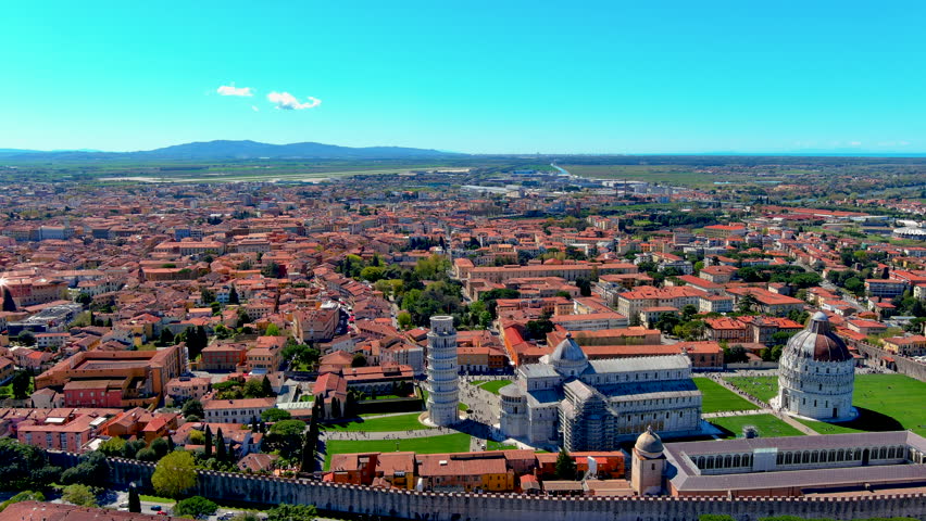 Aerial drone view of Piazza dei Miracoli with the Leaning Tower of Pisa, the cathedral, baptistery and city walls surrounded by red rooftops, Tuscany, Italy, on a sunny day.