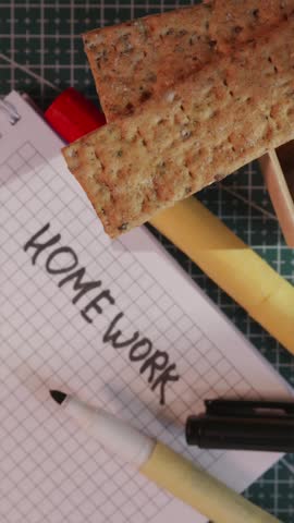 Vertical shot of a hand grabbing a crispbread snack from a plate next to a school notebook labeled Homework on a green cutting mat