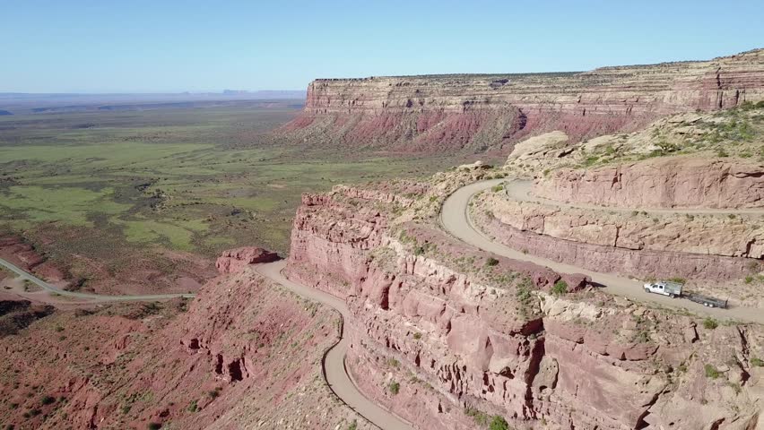 Aerial view showing white pickup truck pulling trailer moving along winding switchbacks on Moki Dugway in Utah red rock landscape with desert terrain under clear blue sky.